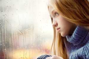 Emotional Portrait of a sad Girl with long blonde Hair. She is looking through a Window in the Rainy Autumn Day