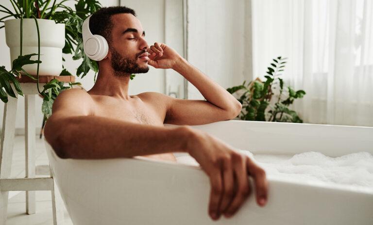Young African-American man in wireless headphones relaxing in bath with foam and enjoying music